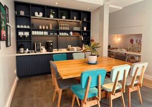 A modern dining area with a wooden table and chairs, a built-in kitchen with shelves, and a potted plant.