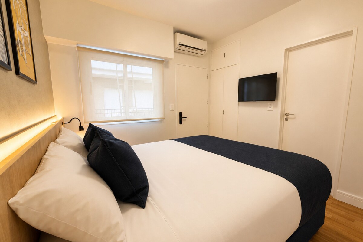 Un dormitorio con cama, ventana con persianas, aire acondicionado, televisor y puerta.