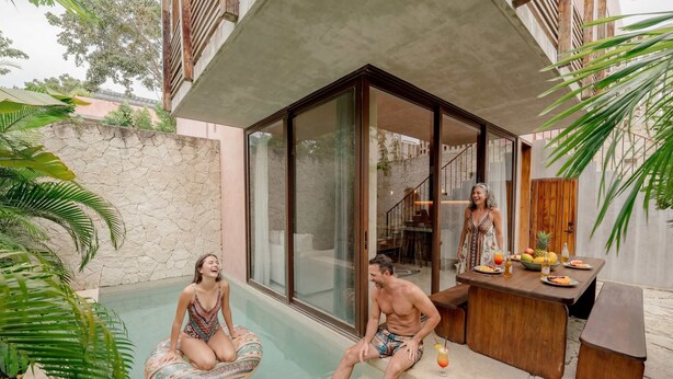 A pool area with a stone wall, a wooden deck, and a dining table with fruit and drinks. - Modern 3BR Villa with 2 Pools & Rooftop Lounge (Tulum)