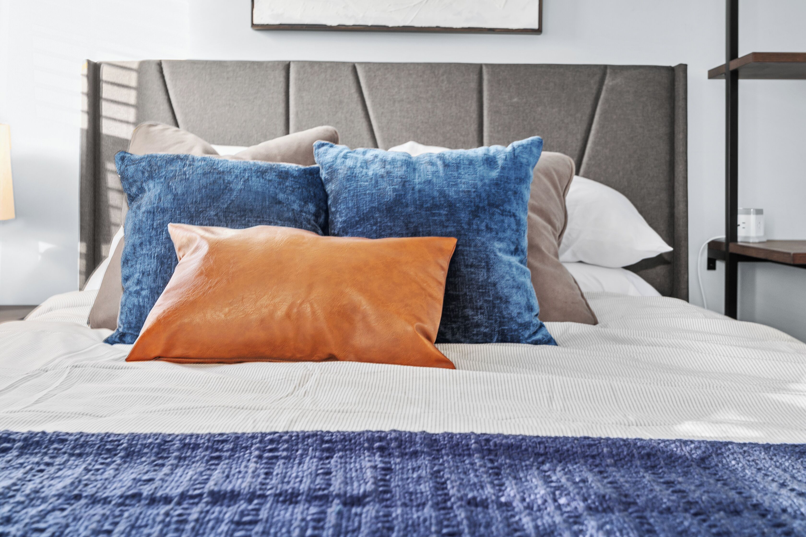 Un lit bien fait, recouvert d’une couverture bleue à texture intéressante, d’un coussin en cuir marron et de plusieurs coussins bleus et beige.