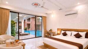 A hotel room with a bed, a desk, a chair, a dining table, and a view of an indoor pool. - OPO Palm Villa Dritan (Udaipur)