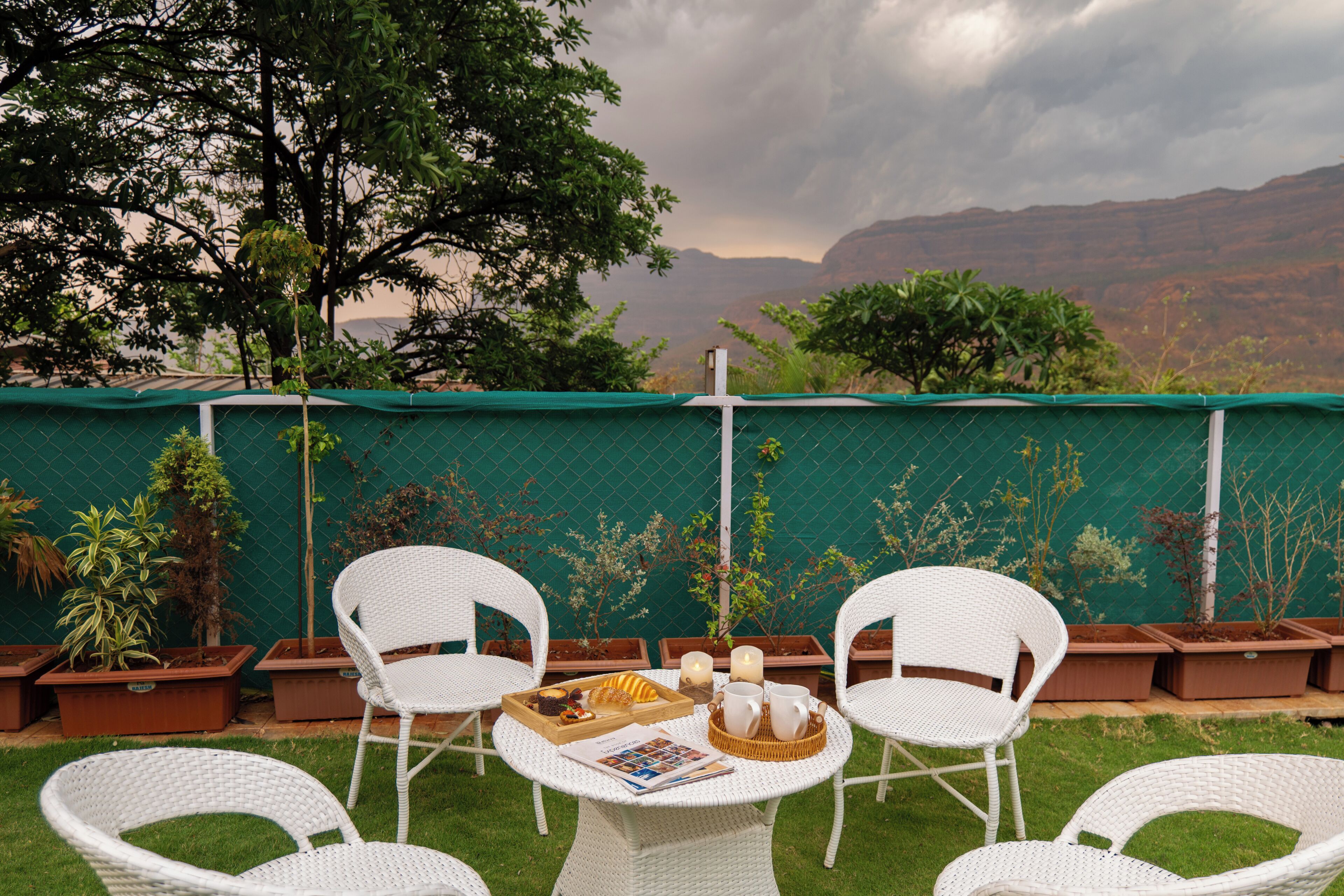 A patio with white wicker chairs, a table with a tray of food and drinks, and potted plants against a green fence with a view of mountains.