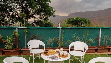 A patio with white wicker chairs, a table with a tray of food and drinks, and potted plants against a green fence with a view of mountains.