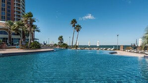A clear blue pool with lounge chairs and palm trees, surrounded by a building with a view of the ocean. - Luxury Portofino Condo | Panoramic Water Views | Pools, Spa & Beach (Pensacola Beach)