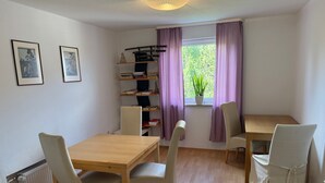 Una habitación con una mesa de madera, dos sillas, un radiador, una ventana con cortinas y estanterías con libros y objetos decorativos.