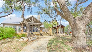 A house with a stone pathway leading to a covered porch, surrounded by large trees and a landscaped garden. - Private 14 Acres | Pool, Pickleball, Hot Tub & Guest House with Views (Wimberley)