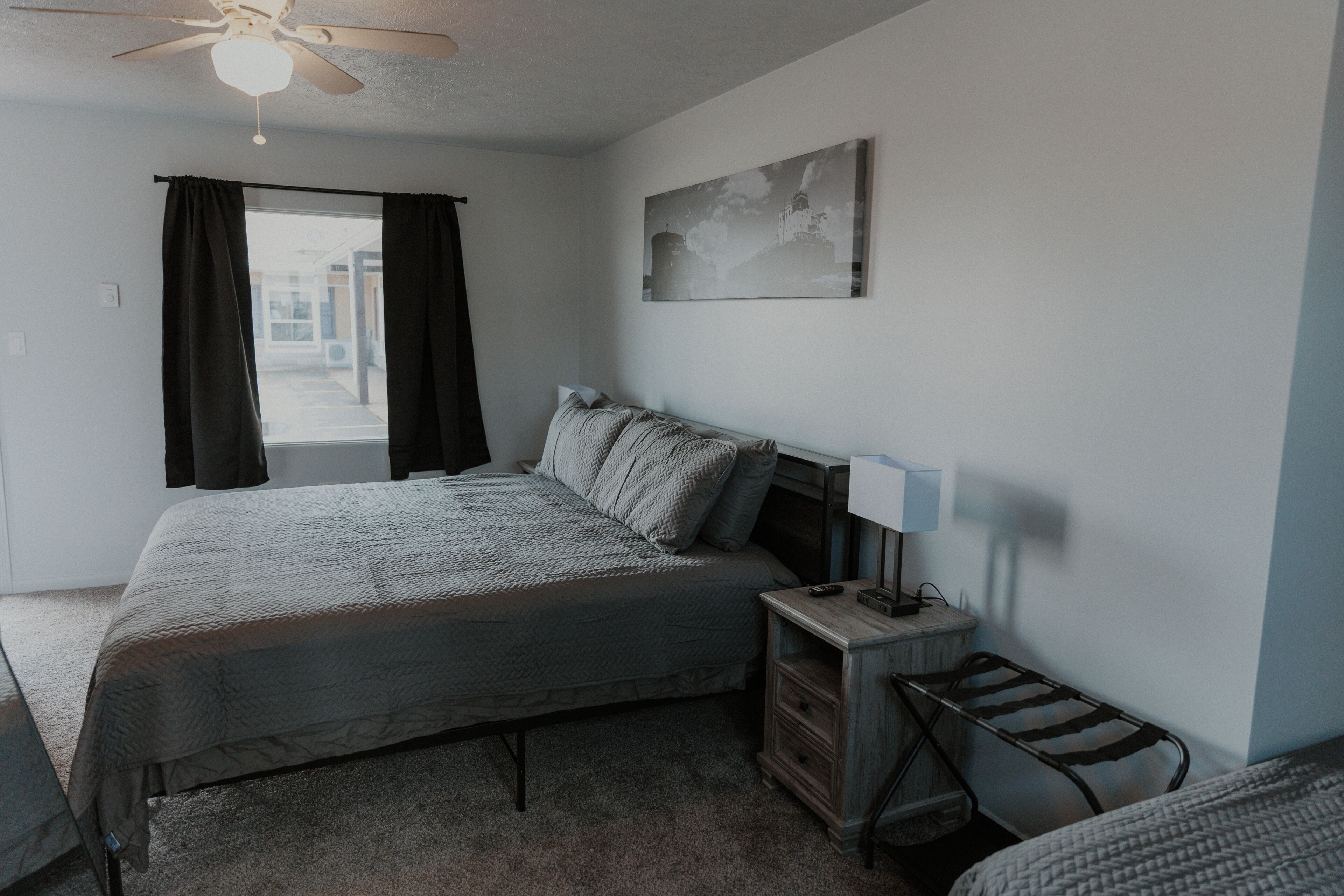 A bedroom with a bed, a nightstand, a lamp, a ceiling fan, and a window with curtains.