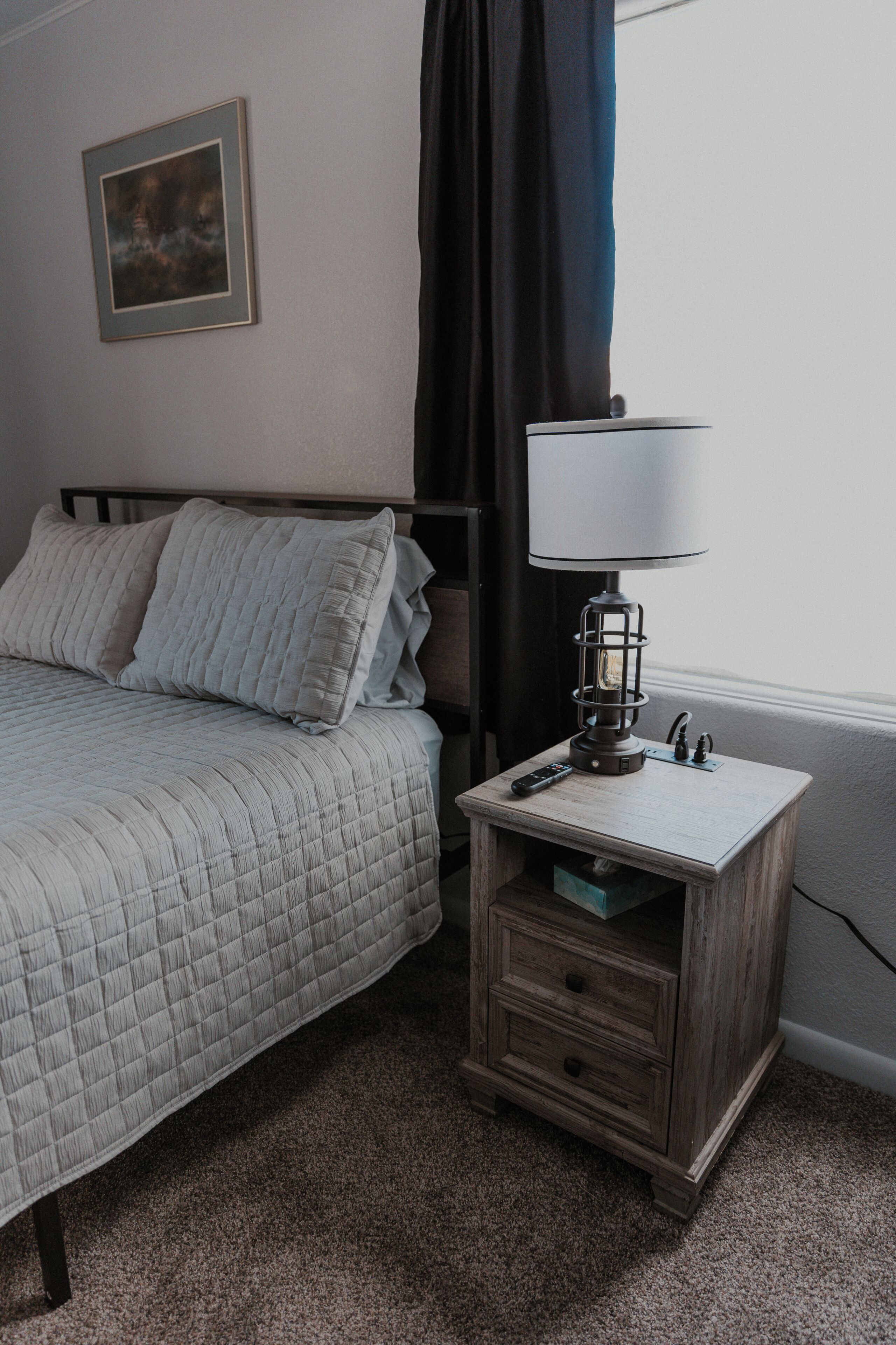 A bedroom with a bed, a bedside table with a lamp, a remote control, and a framed picture on the wall.