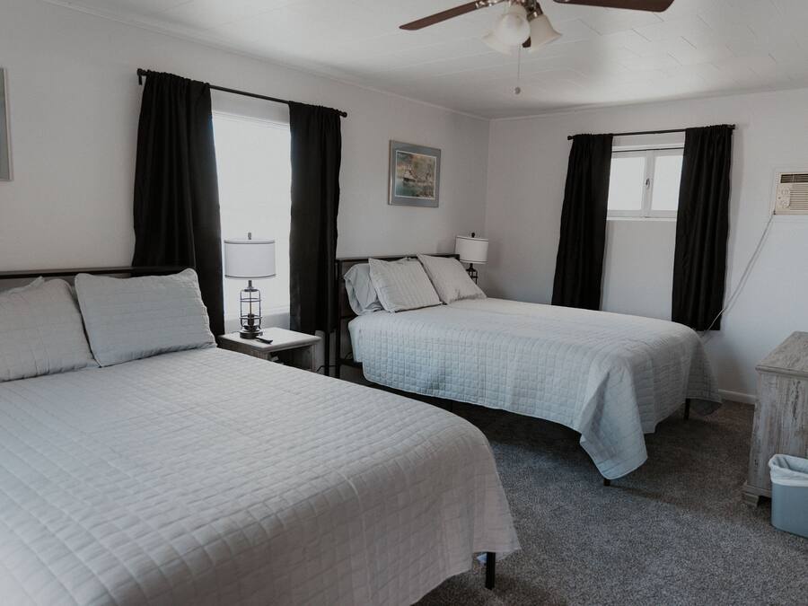 A hotel room with two beds, a ceiling fan, a window with curtains, a nightstand with a lamp, and a small wooden cabinet.
