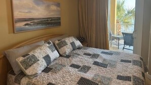 Ett hotellrum med en säng, en målning av ett landskap, en balkong med stolar och utsikt över palmer.