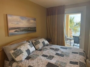 Et hotelværelse med seng, et landskabsmaleri, en balkon med stole og udsigt til palmer.