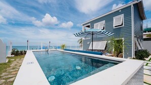 Une piscine située à côté d’une maison à la façade bleue et blanche, avec un patio couvert et une vue sur l’océan.