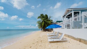 Une plage avec des chaises longues blanches, un parasol bleu et un bâtiment blanc entouré d’une clôture.