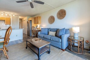 A living room with a sofa, coffee table, and wicker chair. There is a kitchen in the background with cabinets and a refrigerator. - Maui Vista 1208 1 Bedroom Condo (Kihei)