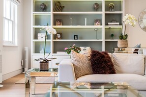 A modern living room with a white sofa, glass coffee table, and a bookshelf filled with decorative items. - Luxury Knightsbridge 4-bedroom House With Central AC (London)