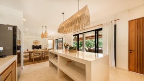 A modern kitchen with a central island, wooden furniture, and a view of greenery outside. - Villa Svara - 4BR Villa w/ Panoramic Ocean Views (Purwakerti)