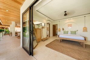 A modern hotel room with a bed, a seating area, a glass door leading to a balcony, and a ceiling fan. - Villa Svara - 4BR Villa w/ Panoramic Ocean Views (Purwakerti)