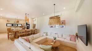 A modern living room with a sectional sofa, a dining area with a wooden table, and a kitchen in the background. - Villa Svara - 4BR Villa w/ Panoramic Ocean Views (Purwakerti)