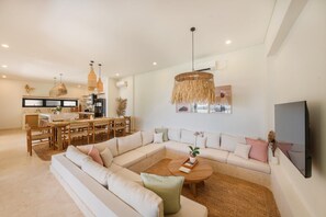 A modern living room with a sectional sofa, a dining area with a wooden table, and a kitchen in the background. - Villa Svara - 4BR Villa w/ Panoramic Ocean Views (Purwakerti)