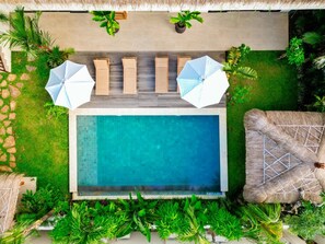 A rooftop pool with lounge chairs and umbrellas, surrounded by lush greenery and a thatched roof.