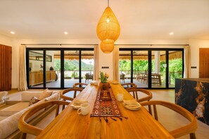 A dining area with a long wooden table, chairs, a hanging light fixture, and large sliding glass doors leading to an outdoor patio with a view of greenery.