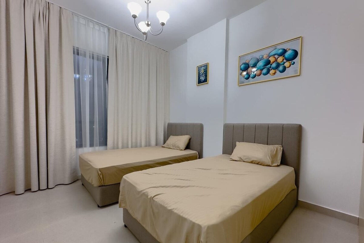 A room with two beds, a large window with curtains, a chandelier, and two framed pictures on the wall.