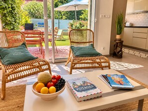 A room with wicker chairs, a table with a bowl of fruit and books, and a view of a garden through a glass door. - Villa Cocoon—Private Pool—Near the Ocean & Montpellier (Mauguio)