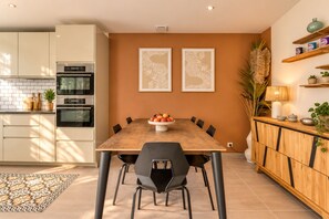 A modern kitchen with white cabinets, a wooden dining table, and black chairs. There are two framed artworks on the wall, a potted plant, and a lamp on the sideboard. - Villa Cocoon—Private Pool—Near the Ocean & Montpellier (Mauguio)