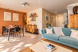 A modern living room with a blue sofa, a dining area with a wooden table and chairs, and a kitchen area with shelves and decorative items. - Villa Cocoon—Private Pool—Near the Ocean & Montpellier (Mauguio)