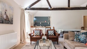 Ein Wohnzimmer mit Glastisch, zwei Ledersesseln, einer Couch und einer Küche im Hintergrund.