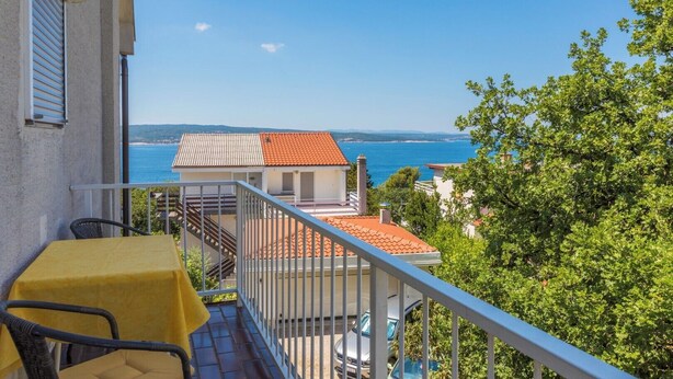 Een balkon met een gele tafel en stoelen, een gebouw met rode dakpannen en uitzicht op zee.