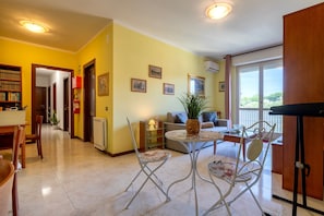 A living room with a sofa, a dining table with chairs, a small round table with a plant, and a kitchen area with cabinets.