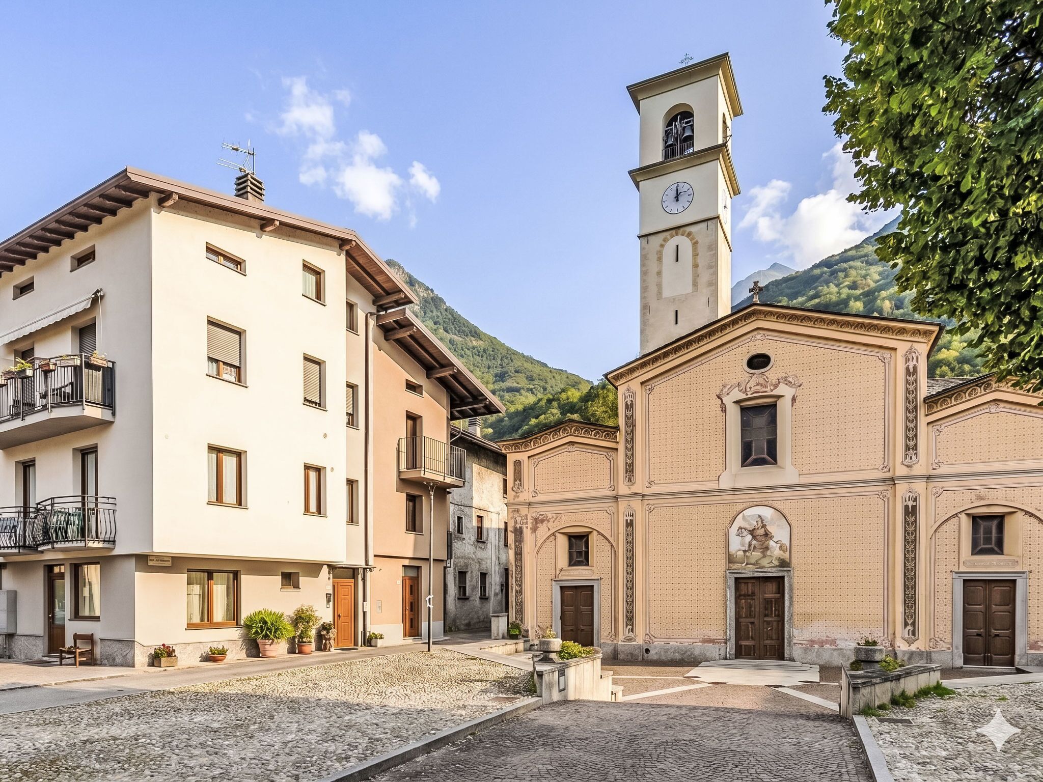 Un edificio histórico con una torre de reloj, un patio con pavimento adoquinado y un telón de fondo montañoso.
