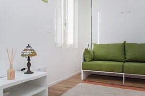 A room with a green sofa, a white table with a lamp, and a window with curtains.