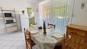 Zona de menjador amb taula per a quatre, cuina amb frigorífic i microones, i vista per una finestra amb cortines.