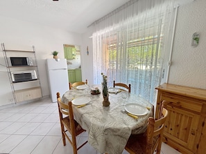 Ein Essbereich mit Tisch für vier Personen, eine Küche mit Kühlschrank und Mikrowelle sowie ein Blick durch ein fenster mit Vorhängen.