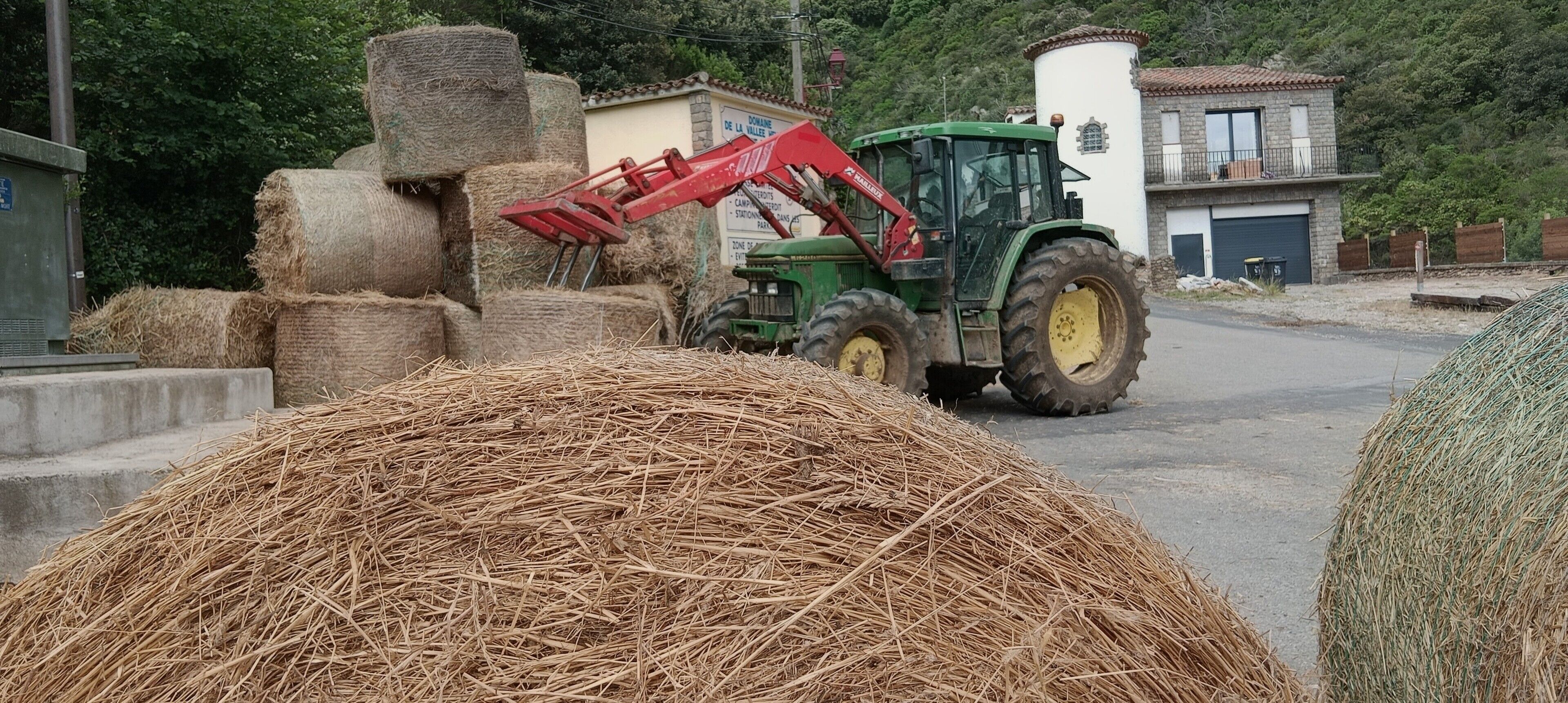 En traktor med rødt redskab, høballer og en bygning med skilt i et landligt område.