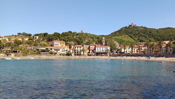 Une ville côtière avec une plage, des bâtiments et une colline surmontée d’un moulin à vent.