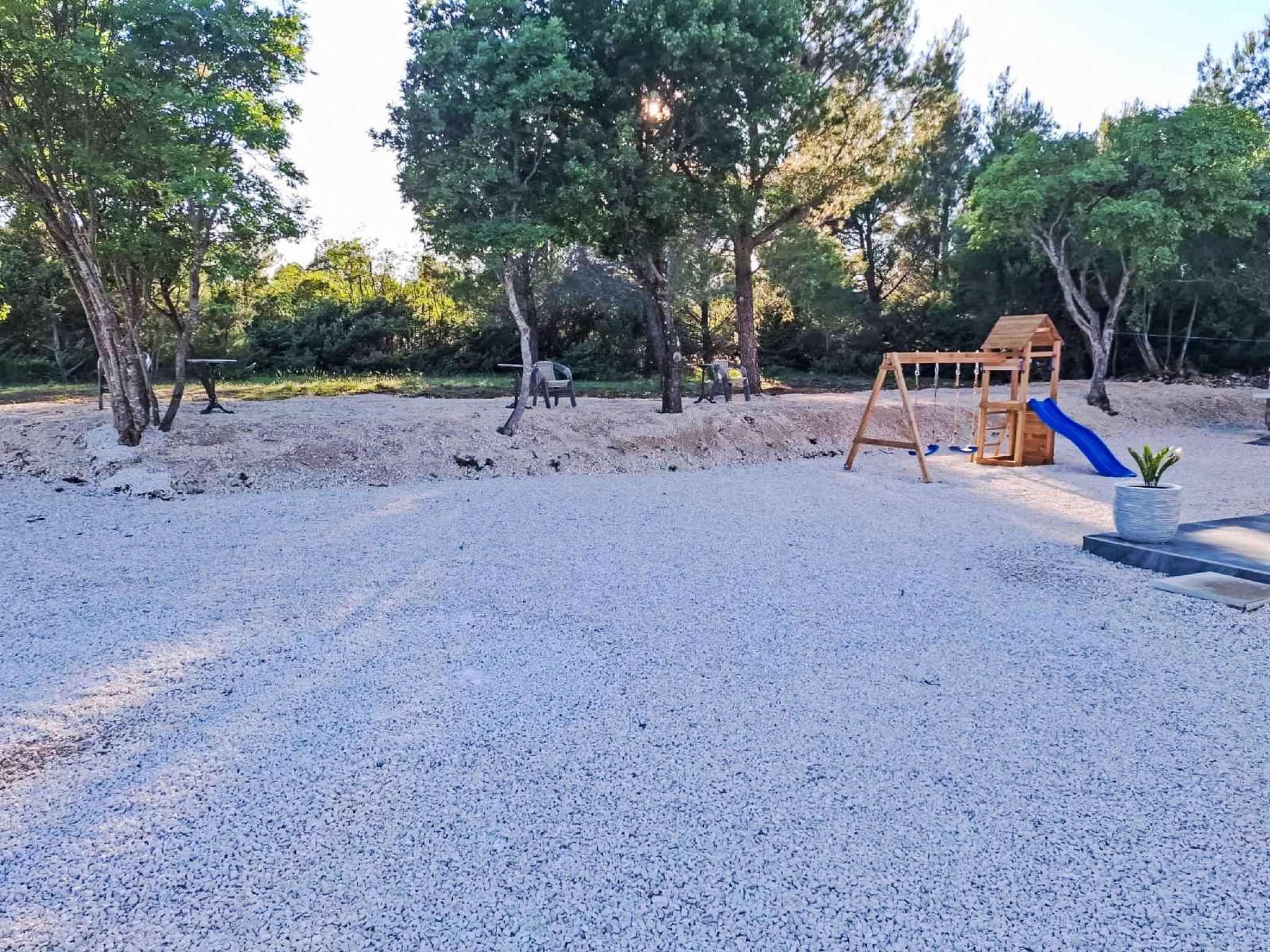 Ein Spielplatz mit Schaukel und Rutsche, umgeben von Bäumen und einem Kiesbereich.
