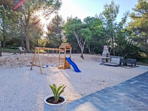 Ein Spielplatz mit Schaukel und Rutsche, umgeben von Bäumen und einer Grillgelegenheit.