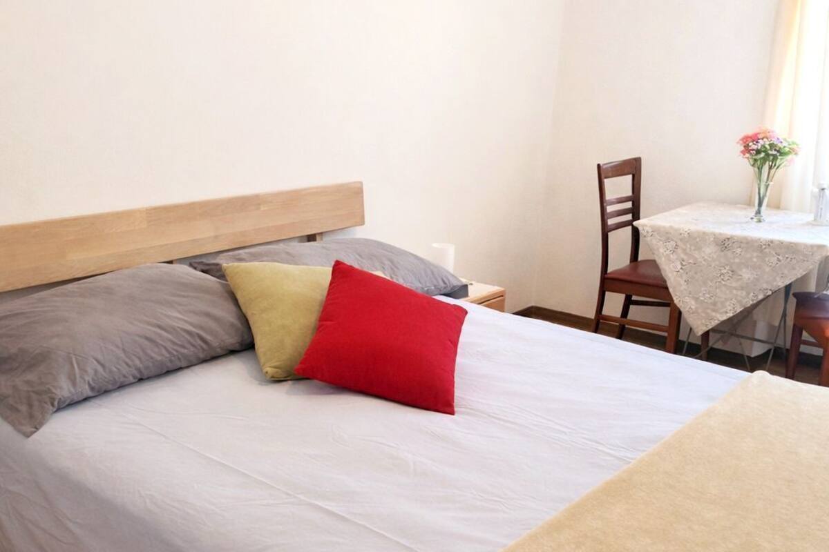 Ein Bett mit einem Holz-Kopfteil, einer grau-beigen Decke und einem roten Kopfkissen.