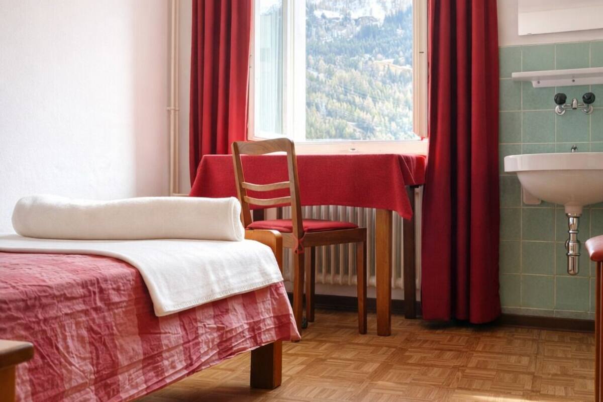 Ein Zimmer mit Bett, Tisch mit Stuhl, Fenster mit Vorhängen und einem Waschbecken.