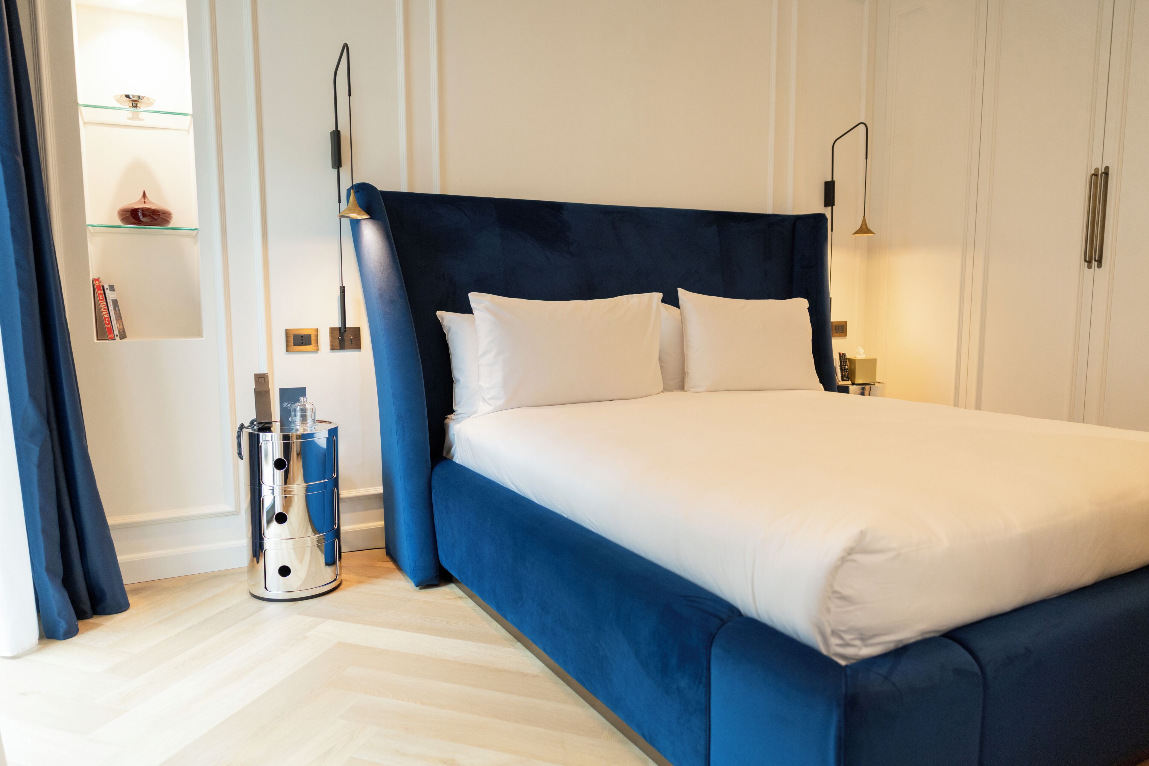 A neatly made bed with a dark headboard, a bedside table with a lamp, and a small shelf with books and decorative items.