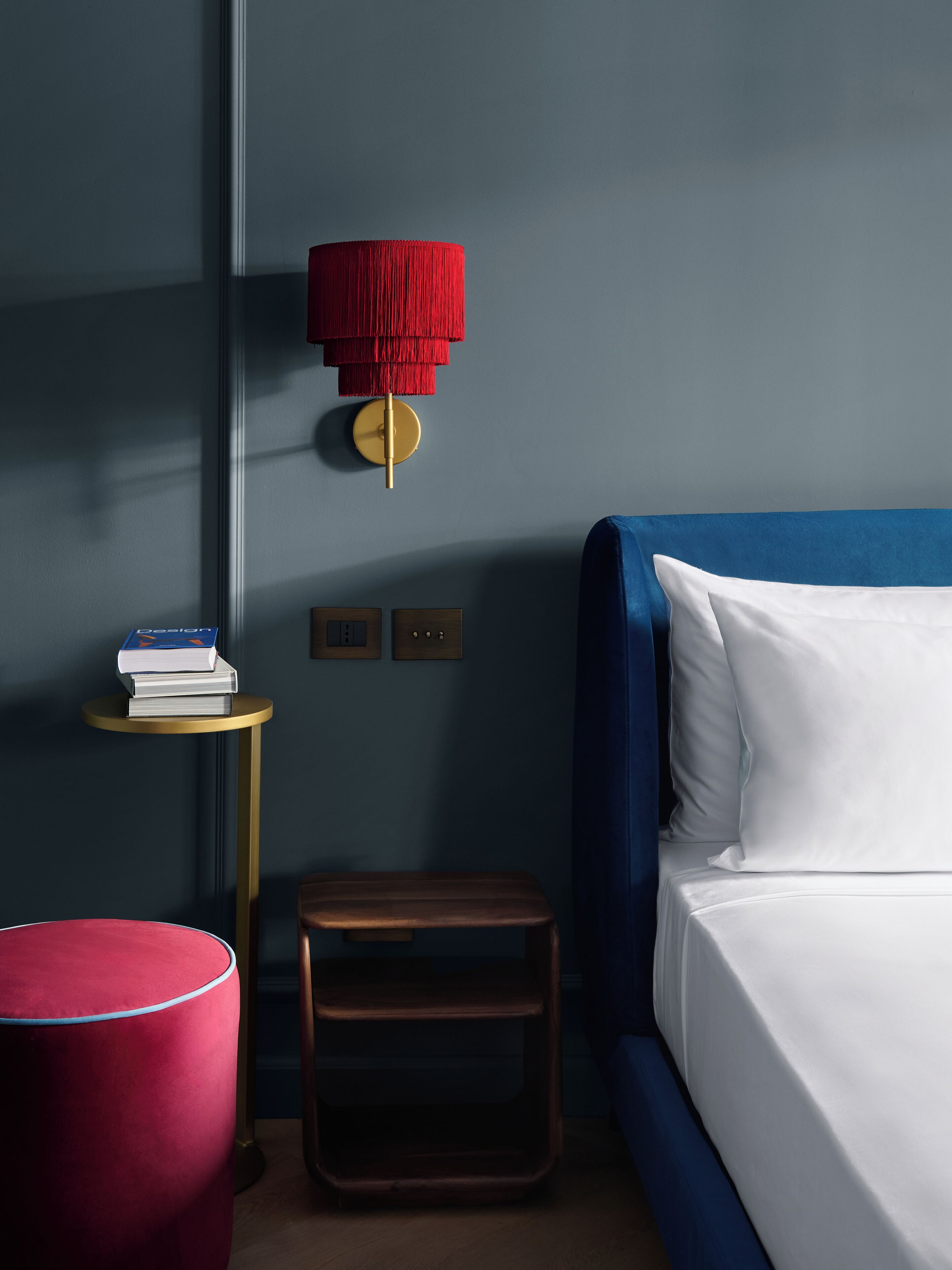 Una camera da letto con un letto, un comodino, un poggiapiedi rosso e una lampada a parete.