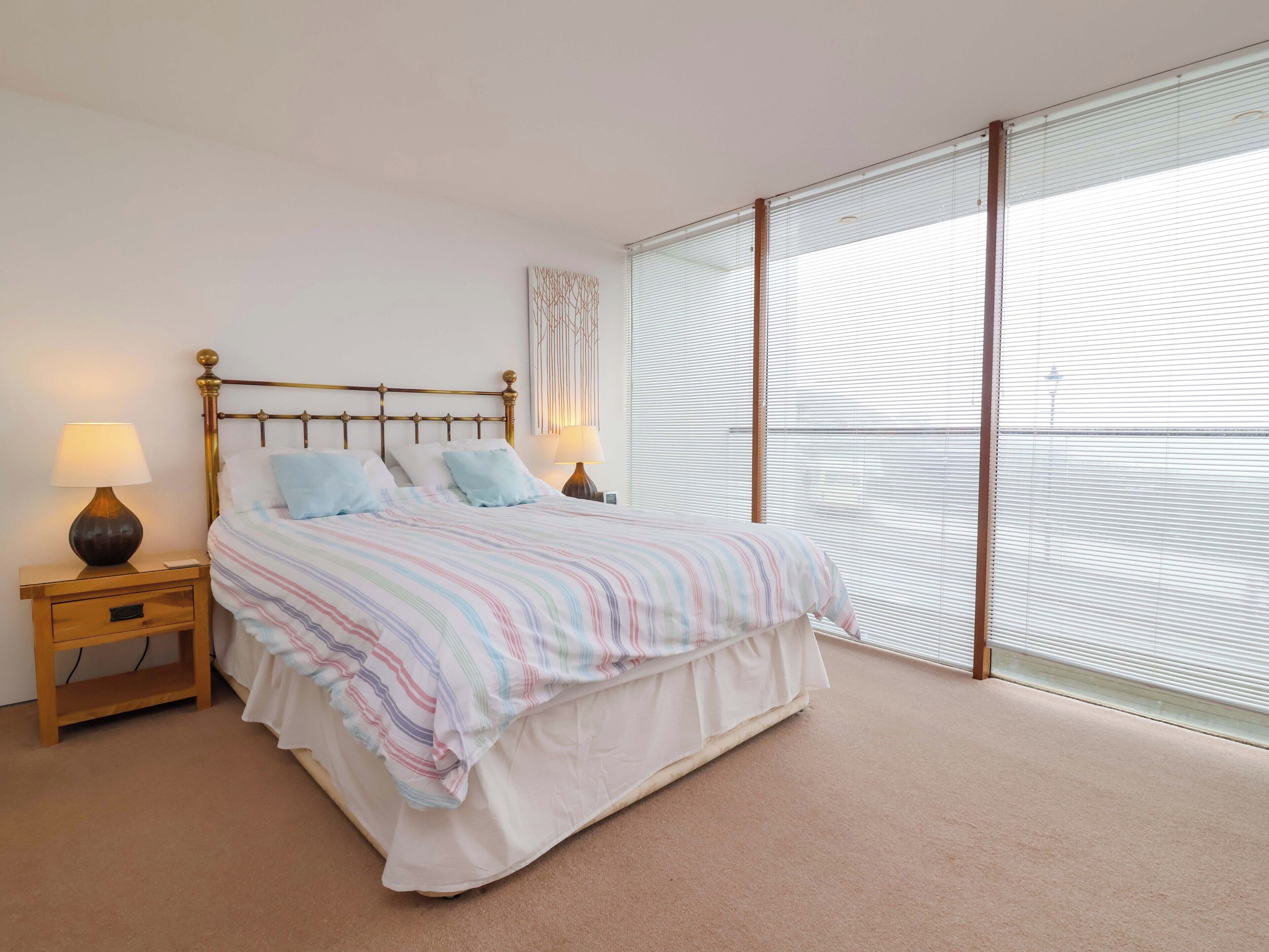 A bedroom with a bed, nightstands, a lamp, and a large window with blinds.