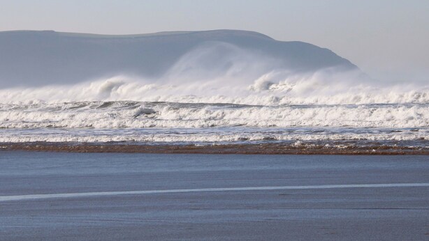 A beach with waves crashing onto the shore and a distant mountain. - 12 Nautilus (Bideford)