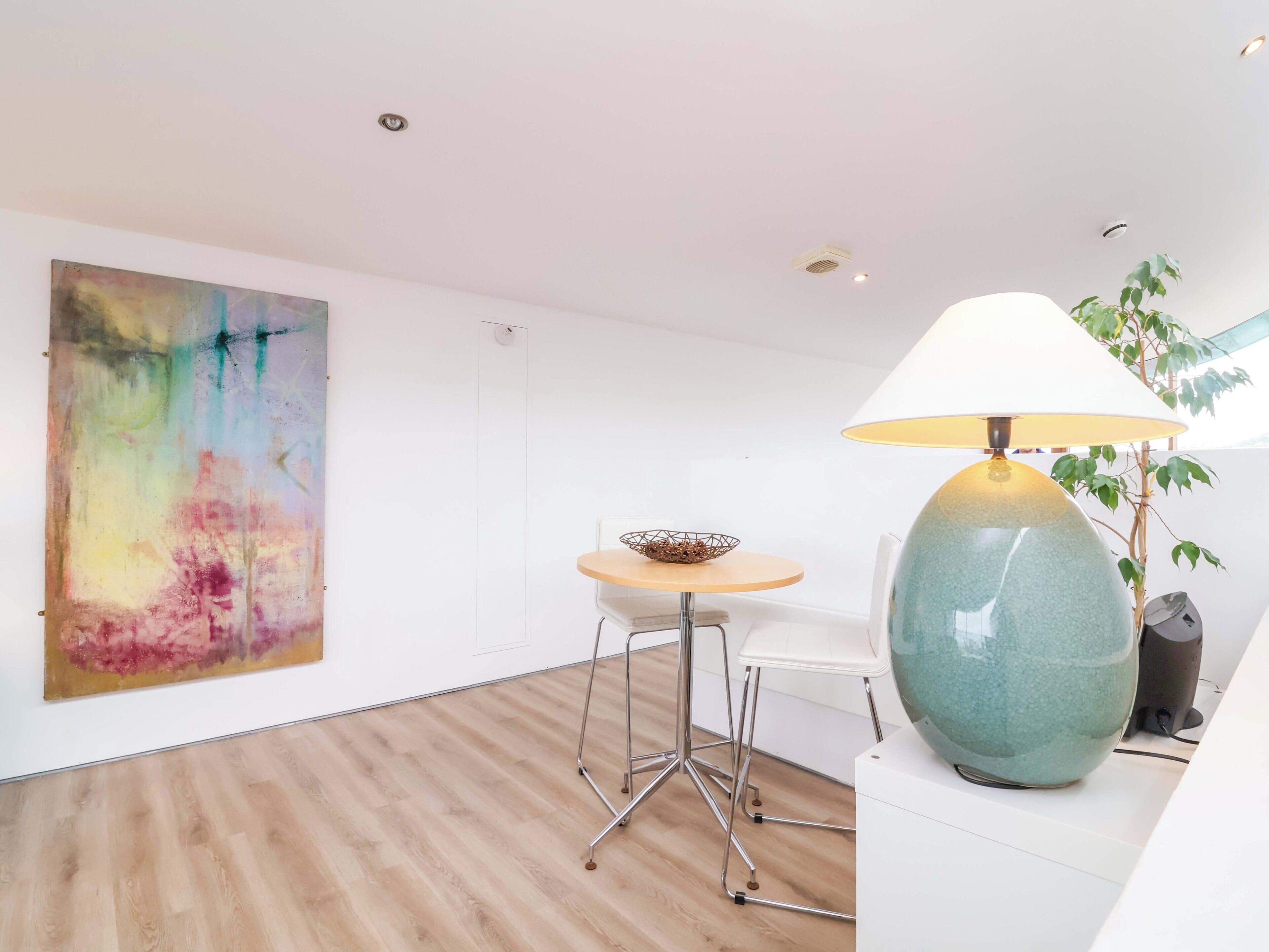 A modern room with a wooden floor, a round table with a bowl, a white chair, a large abstract painting, and a green lamp.