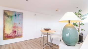 A modern room with a wooden floor, a round table with a bowl, a white chair, a large abstract painting, and a green lamp.