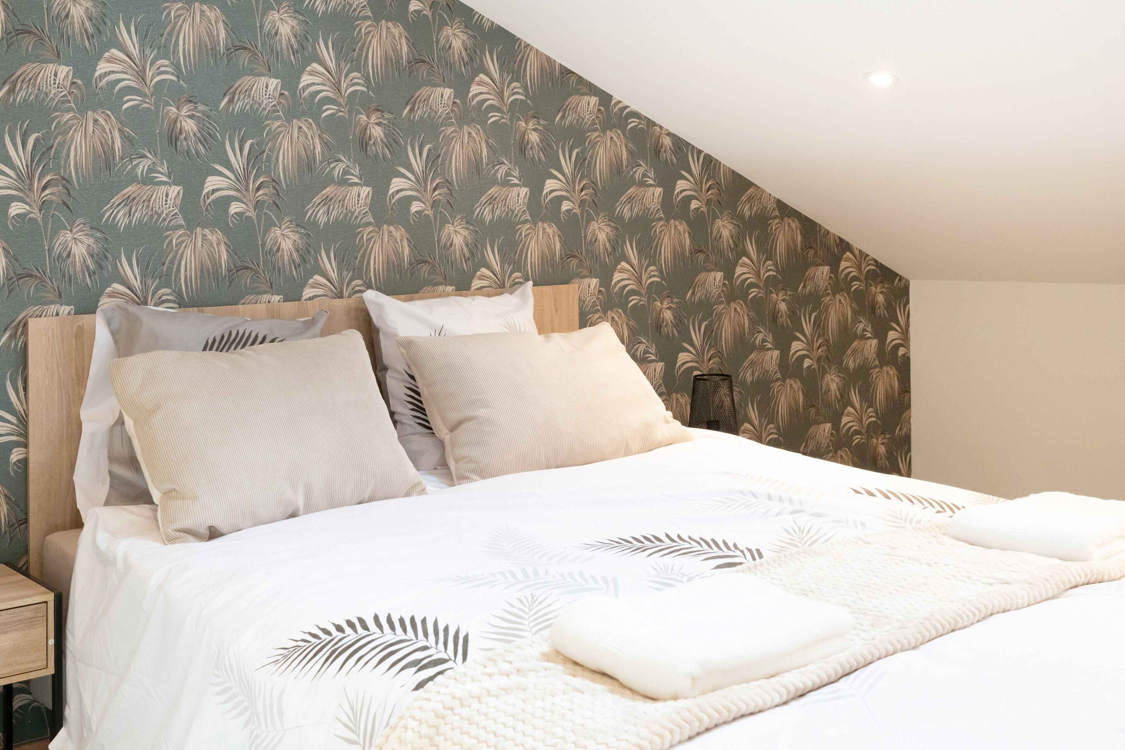 A neatly made bed with a leaf-patterned bedspread, a wooden headboard, and a wall with a leaf-patterned wallpaper.