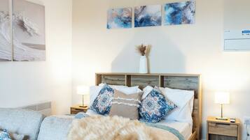 Een slaapkamer met een bed, nachtkastjes, een lamp, een vaas met gedroogde planten en abstracte wanddecoratie.
